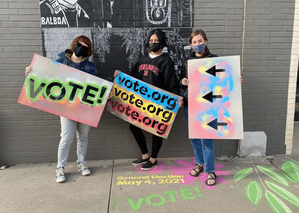 Three people stand in a line holding signs that read "Vote!"