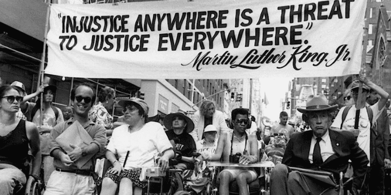 A black and white photo shows disability justice pioneer, Judy Heumann, protesting on Madison Avenue, in Manhattan, New York in 1970 alongside other proud members of Disabled In Action. Above their heads is a huge white banner that reads, “Injustice anywhere is a threat to justice everywhere.” in black text.