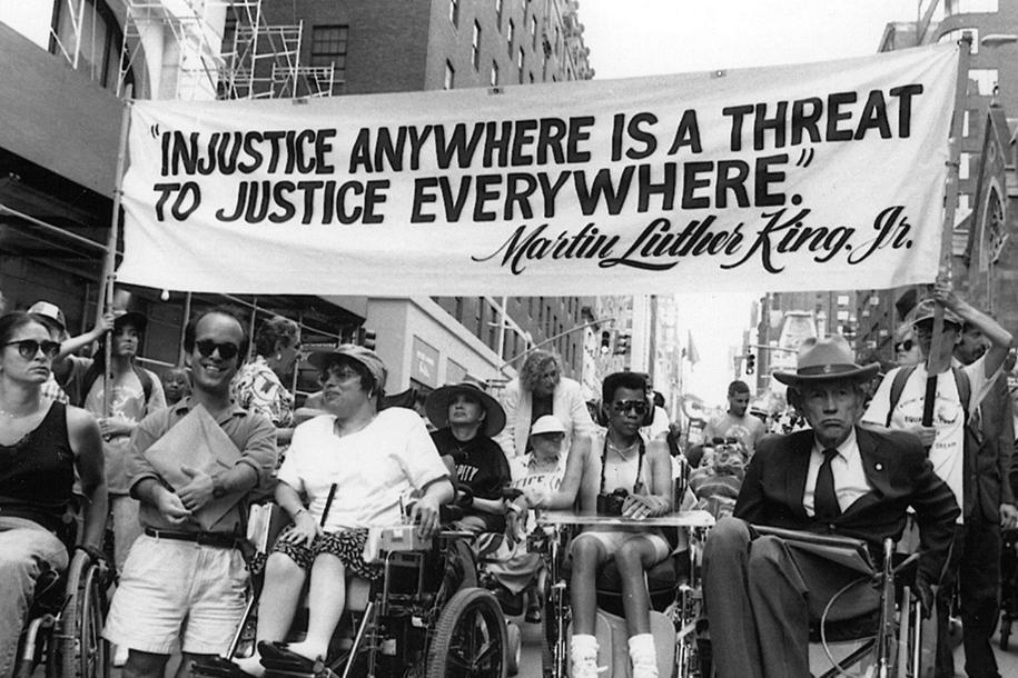 A black and white photo shows Judy Heumann, protesting on Madison Avenue, in Manhattan, New York in 1970 alongside other proud members of Disabled In Action. Above their heads is a LARGE white banner that reads, “Injustice anywhere is a threat to justice everywhere.” in black text.