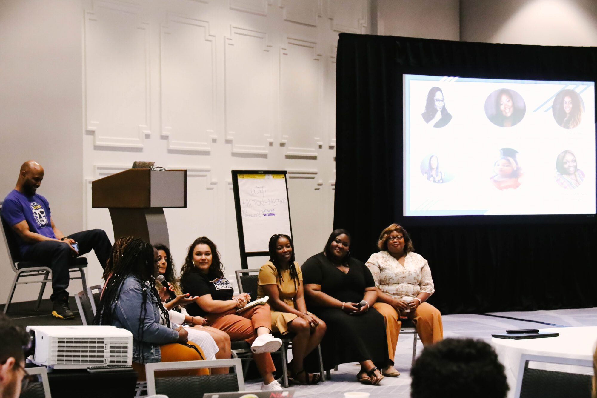 A seated panel discussion is highlighted featuring five women of color sat at the front of a stage.