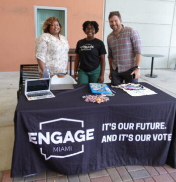 Three participants from the 2023 Data Convening in Miami, Florida stand smiling behind a table with a dark blue covering with white text that reads, "Engage Miami, It's our future, and it's our vote". There are a few pamphlets other event materials spread across the table to greet guests.