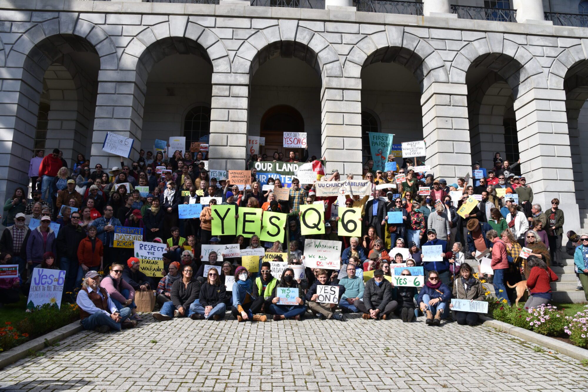 A large crowd of protesters pose for a group photo at a rally at the Maine State House on Indigenous Peoples Day, 2023. Many people are holding signs that say "Yes On 6". 
