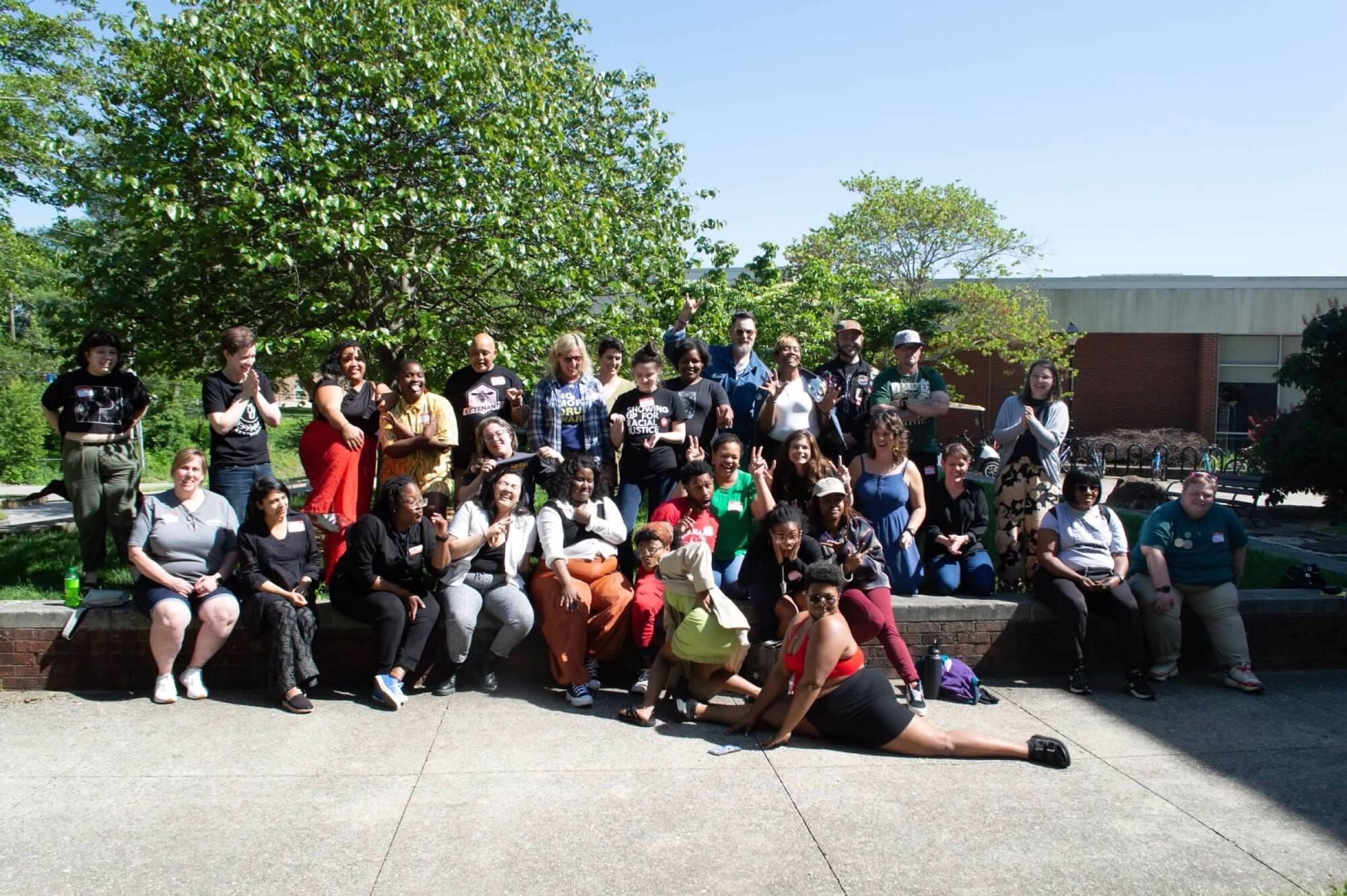 Members of the Kentucky Civic Engagement Table pose together for a group photo in front of trees outside. People are seen smiling and laughing. One member towards the bottom of the photo is doing a split pose on the ground. 