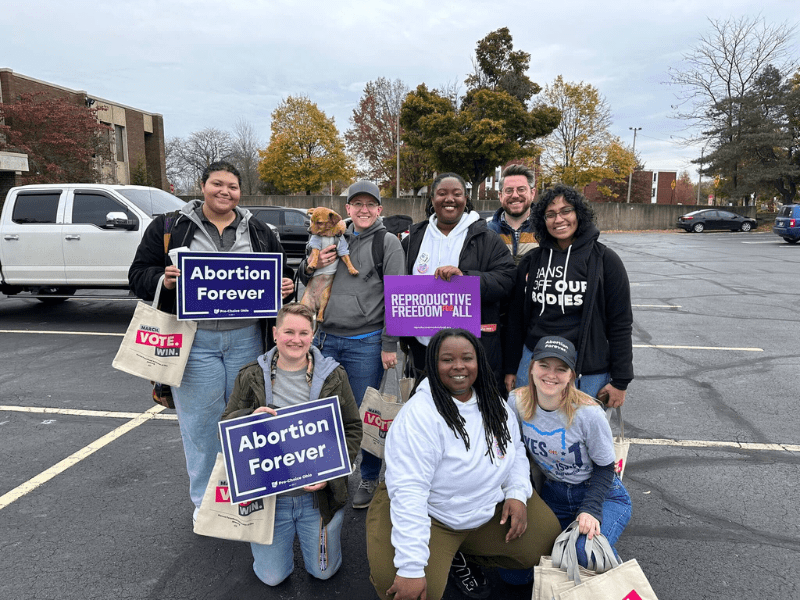 Members of Pro Choice Ohio, Planned Parenthood Advocates of Ohio and Ohioans United for Reproductive Rights post for a group photo in a parking lot. A racially diverse group of people are seen smiling and holding signs that say, "Reproductive Freedom For All". 