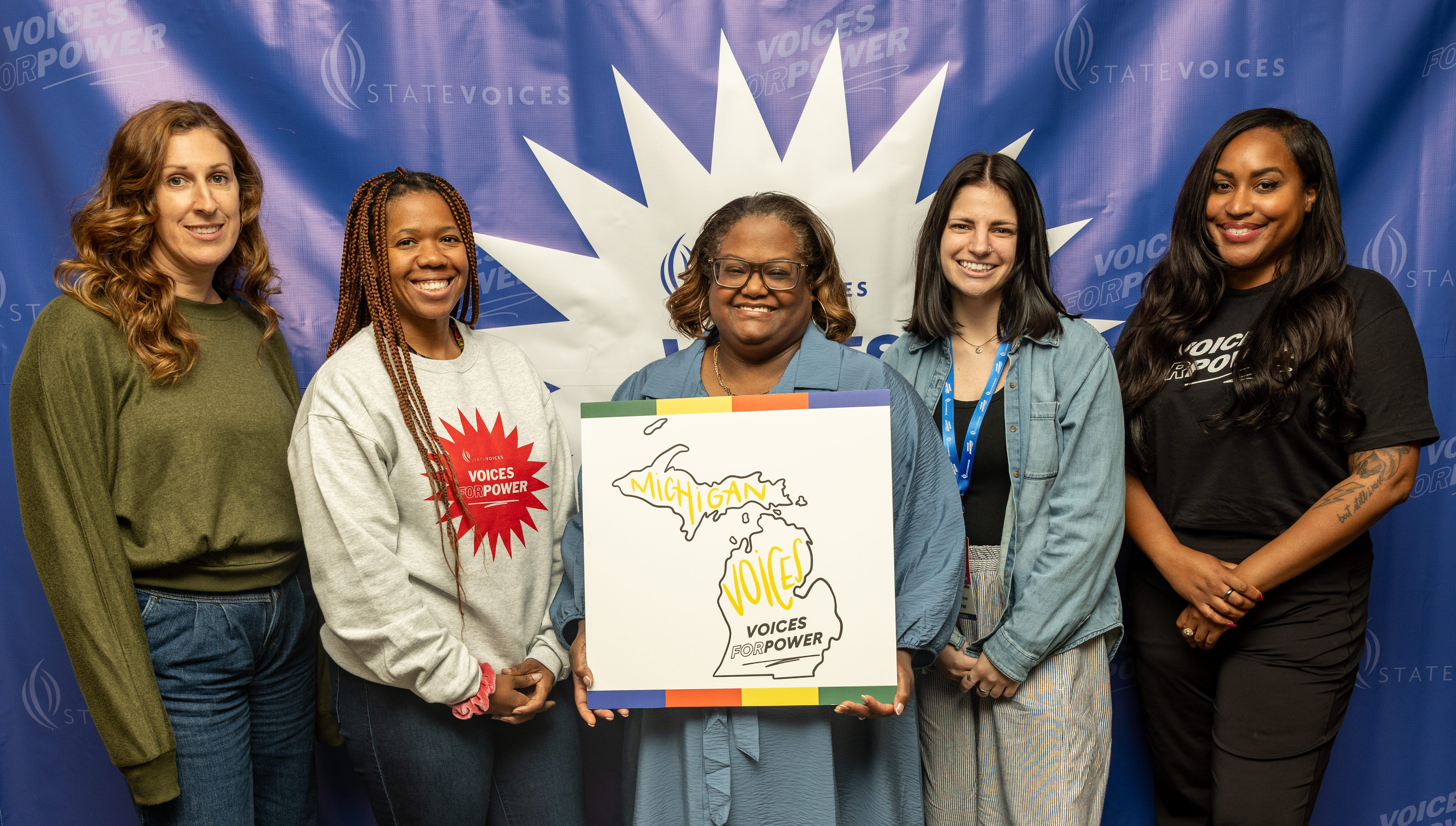 Advocates with Michigan Voices smile and hold up a map of their state that reads "Michigan Voices. Voices for Power."